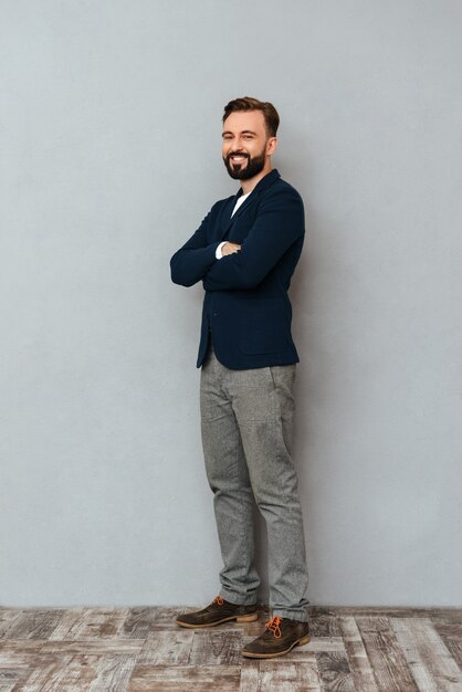 Image pleine longueur d'homme barbu en vêtements de travail posant avec les bras croisés et regardant la caméra sur gris