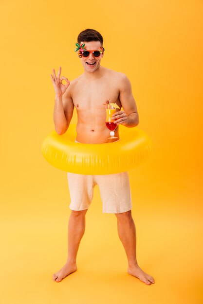 Image pleine longueur d'un homme nu souriant en short, des lunettes de soleil inhabituelles et un anneau gonflable tenant un cocktail tout en montrant un signe ok