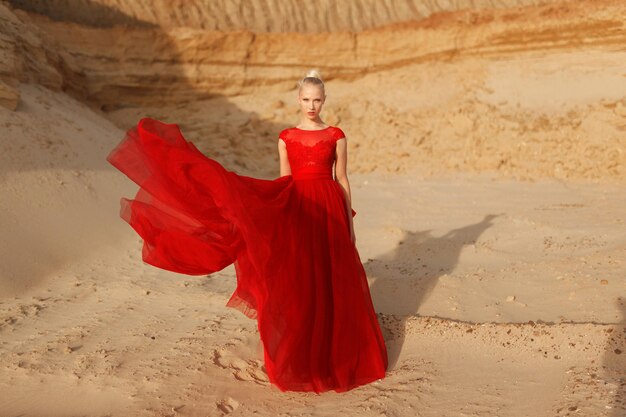 Image pleine longueur d'une jeune femme blonde en robe rouge avec tissu volant sur fond de sable doré.