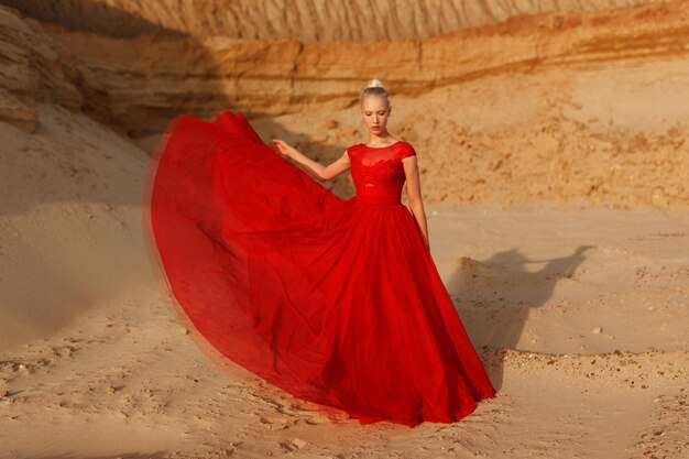 Image pleine longueur d'une jeune femme sensuelle en robe ondulante rouge avec un tissu volant sur fond de sable doré.