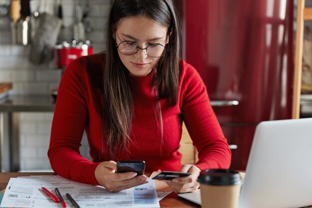 Image recadrée d'une femme brune aux taches de rousseur dans des vêtements décontractés rouges, détient un téléphone intelligent moderne et une carte en plastique dans les mains