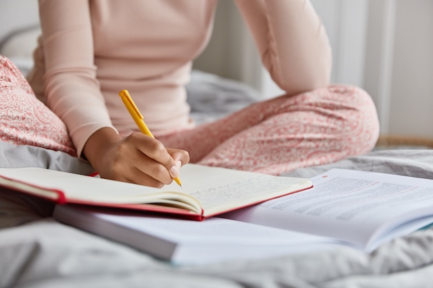 Image recadrée d'une femme méconnaissable en chemise de nuit, écrit des informations dans le bloc-notes, réécrit le sujet du manuel, pose seule au lit, a une belle écriture. Gros plan, se concentrer sur l'écriture