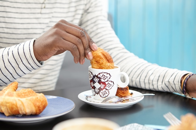 Photo gratuite image recadrée d'un homme afro-américain méconnaissable trempant un croissant dans une tasse de cappuccino, savourant un délicieux petit-déjeuner seul au café, assis à table avec une tasse et une pâtisserie. effet de film