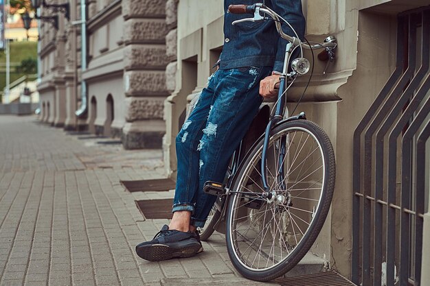 Image recadrée d'un homme à la mode dans des vêtements élégants appuyé contre un mur avec un vélo de ville dans une rue.