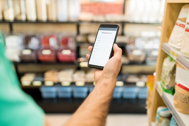 Image recadrée d'un jeune homme vérifiant la liste de courses dans un smartphone à l'épicerie