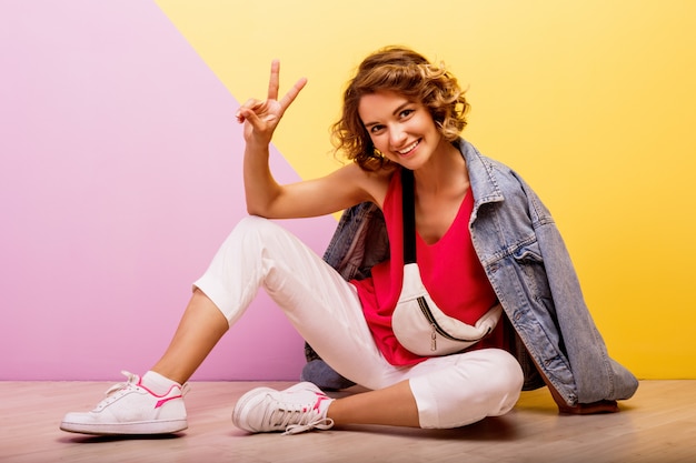 Image Studio de femme brune souriante belle portant une tenue sportive élégante et une veste en jean assis sur le sol.