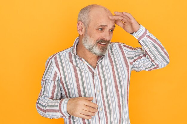 Image Studio de frustré et perplexe senior homme mal rasé chauve tenant la paume sur le front pour protéger les yeux du soleil, à la recherche de poussière ou à la recherche de quelque chose.