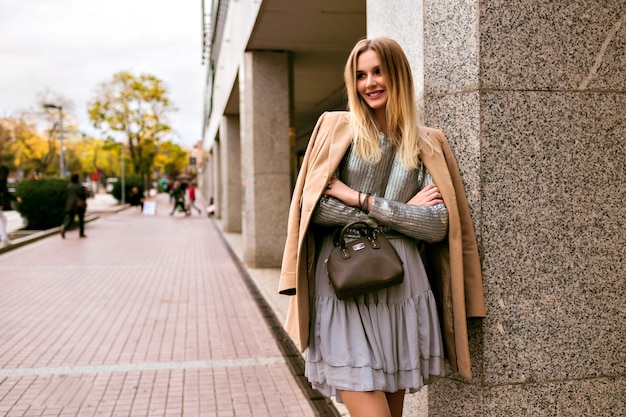 Image de style de rue de mode d'une femme élégante blonde, vêtue d'une robe en soie de luxe, d'un pull à la mode, d'un manteau en cachemire et d'un sac en cuir, des couleurs douces et chaudes, une ambiance printemps automne mi-saison.