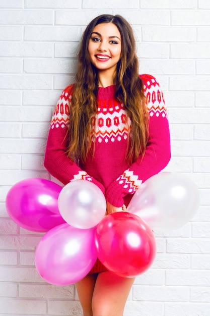 Image de style de vie intérieur d'une jolie fille brune drôle avec un maquillage lumineux et des cheveux longs, portant un pull à la mode et tenant des ballons de fête roses.