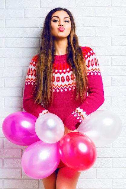 Image de style de vie intérieur d'une jolie fille brune drôle avec un maquillage lumineux et des cheveux longs, portant un pull à la mode et tenant des ballons de fête roses.