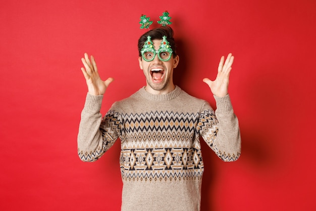 Photo gratuite image d'un type caucasien excité et émerveillé dans des lunettes de fête, pull de noël, levant les mains et faisant une grande annonce, profitant de la célébration du nouvel an, fond rouge.