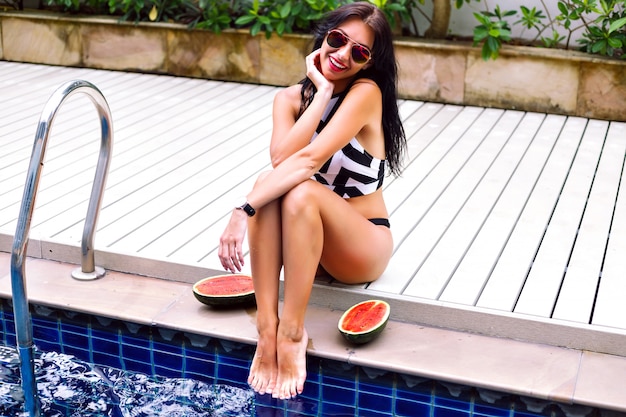 Image de vacances de style de vie d'été en plein air d'une femme sexy élégante, posant près de la piscine à ses vacances, bikini de style géométrique de mariage et lunettes de soleil, mettre des pastèques autour, des émotions heureuses.