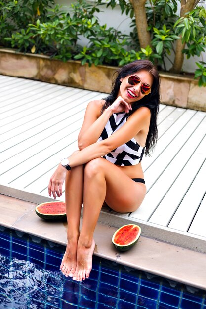Image de vacances de style de vie d'été en plein air d'une femme sexy élégante, posant près de la piscine à ses vacances, bikini de style géométrique de mariage et lunettes de soleil, mettre des pastèques autour, des émotions heureuses.