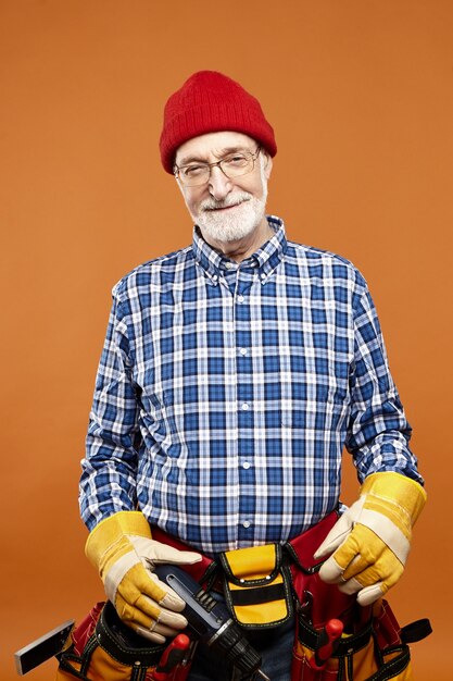 Image verticale de l'heureux constructeur masculin âgé expérimenté avec barbe grise posant au mur blanc, portant des lunettes, des gants en caoutchouc, un chapeau et une ceinture avec des instruments, à la recherche d'un large sourire