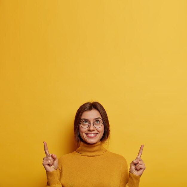 Image verticale de jolie femme souriante pointe l'index ci-dessus, se vante de son nouveau plafond, porte des turtlenck occasionnels