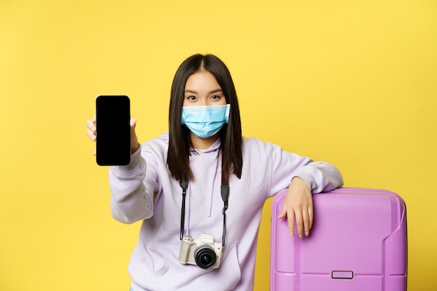 Image d'une voyageuse, une touriste asiatique montre l'écran de son téléphone, un passeport de santé covid sur une application pour smartphone, portant un masque médical et une valise, fond jaune.