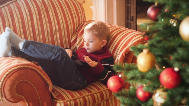 Images 4k d'un petit garçon allongé sur un canapé dans le salon et utilisant une tablette tactile. enfant s'amusant pendant les vacances d'hiver et les célébrations.
