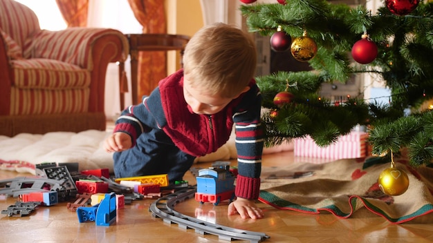 Images k de petit garçon construisant un chemin de fer jouet autour de l'arbre de noël dans le salon. enfant recevant des cadeaux et des cadeaux pendant les vacances d'hiver et les célébrations