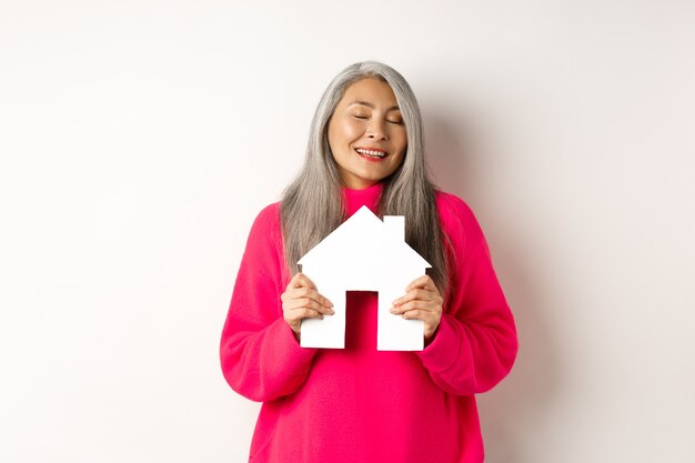 Immobilier. Belle dame asiatique rêveuse étreignant le modèle de maison de papier avec les yeux fermés, souriant comme rêvant d'acheter un appartement, debout sur fond blanc