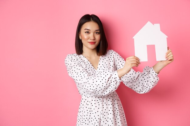 Immobilier belle femme asiatique démontrant un modèle de maison en papier regardant une annonce confiante de caméra...