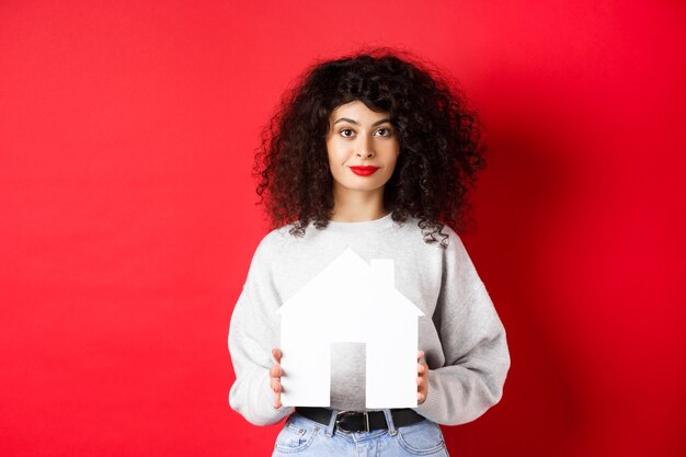 Immobilier. Jeune femme de race blanche en vêtements décontractés montrant la découpe de la maison en papier, l'achat d'une propriété ou la location d'un appartement, debout sur fond rouge