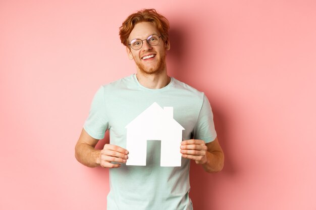 Immobilier. Jeune homme gai aux cheveux rouges, portant des lunettes et un t-shirt, montrant la découpe de la maison en papier et souriant, achetant un appartement, fond rose.