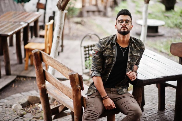 Impressionnant beau grand homme macho à la barbe arabe dans des verres et une veste militaire assis à une table en bois extérieure du restaurant