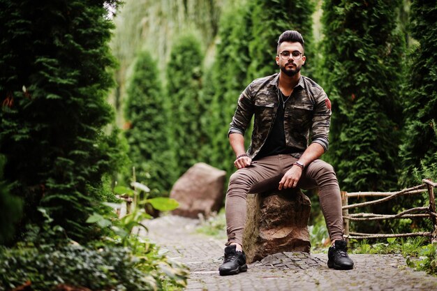 Impressionnant beau grand homme macho barbe arabe à lunettes et veste militaire posé en plein air assis sur la pierre