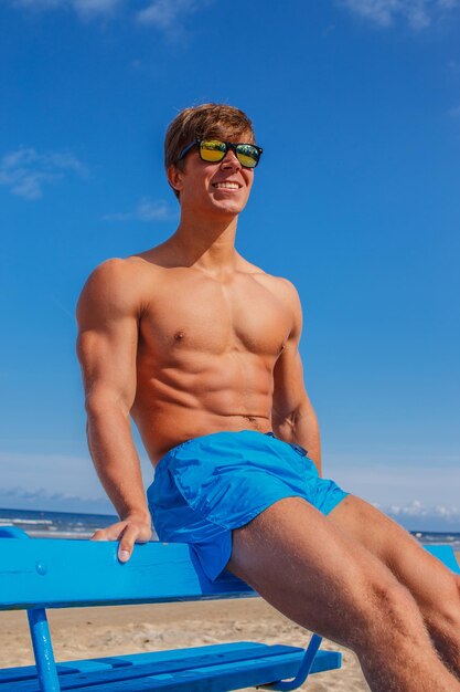 Impressionnant jeune homme musclé en short de bain bleu assis sur un banc sur le ciel bleu et la mer.