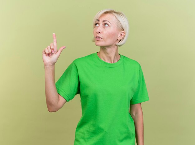 Impressionné d'âge moyen blonde femme slave tournant la tête à l'autre à la recherche et pointant vers le haut isolé sur mur vert olive avec espace copie