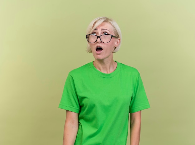 Impressionné d'âge moyen femme slave blonde portant des lunettes à la recherche d'isolement sur le mur vert olive avec espace copie