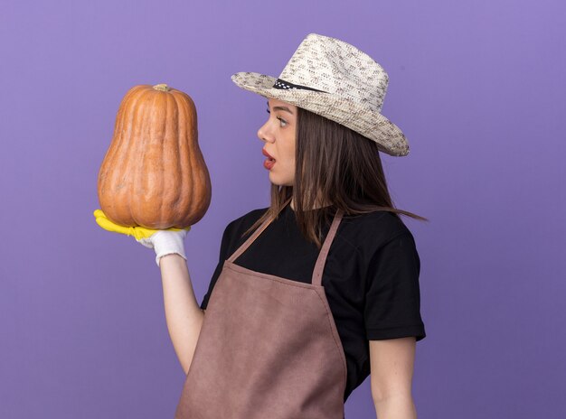 Impressionné assez caucasien femme jardinier portant chapeau de jardinage et des gants tenant et regardant la citrouille sur violet