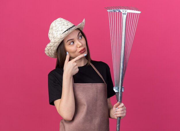 Impressionné assez caucasien femme jardinier portant chapeau de jardinage tenant et pointant sur le râteau à feuilles sur rose