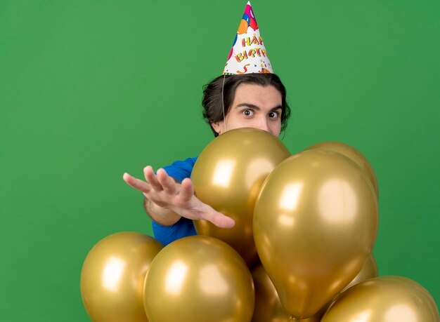 Impressionné bel homme portant une casquette d'anniversaire se dresse avec des ballons d'hélium étendant les mains isolés sur le mur vert