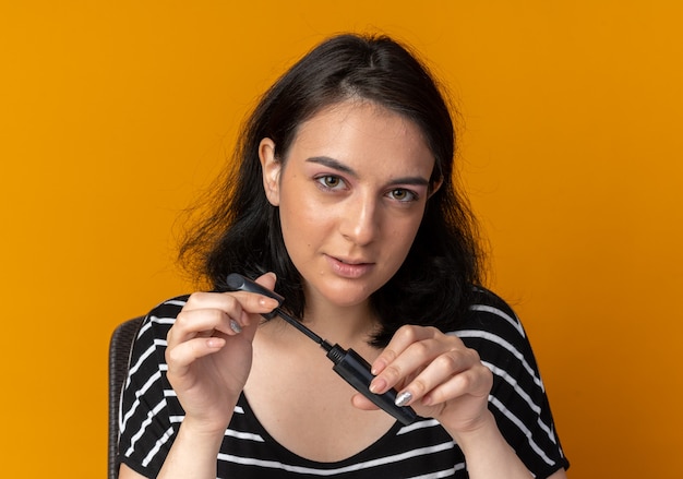 Impressionné à la belle jeune fille tenant un mascara isolé sur un mur orange