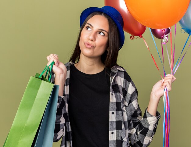 Impressionné à côté de la belle jeune fille portant un chapeau bleu tenant des ballons avec un sac-cadeau