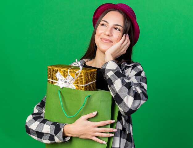 Impressionné à côté de belle jeune fille portant un chapeau de fête tenant un sac-cadeau mettant la main sur la joue