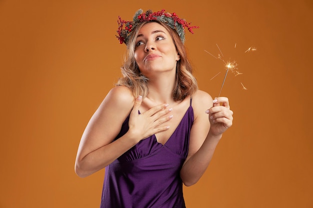 Impressionné à côté de la belle jeune fille vêtue d'une robe violette avec une couronne tenant des cierges magiques mettant la main elle-même isolée sur fond marron