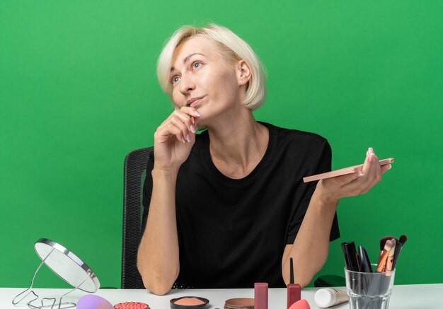 Impressionné du côté de la belle jeune fille assise à table avec des outils de maquillage tenant une palette de fards à paupières avec un pinceau de maquillage isolé sur un mur vert