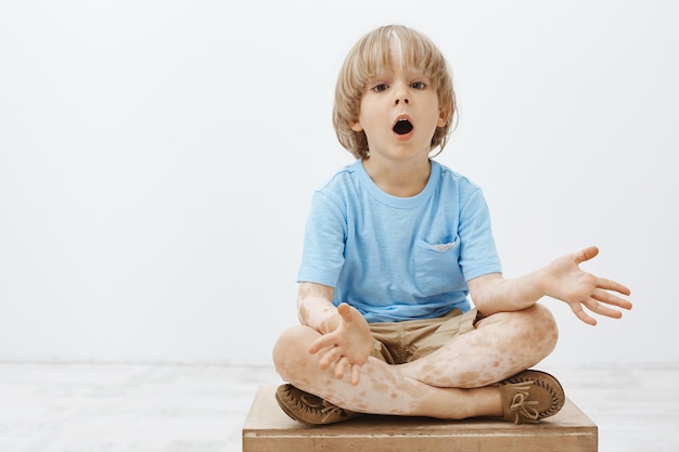 impressionné enfant blond curieux avec vitiligo assis avec les pieds croisés et la mâchoire tombante, écartant les paumes