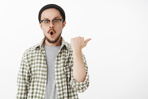 Impressionné et étonné de beau mec avec une barbe dans des lunettes et un bonnet pliant les lèvres dans un son wow pointant à droite avec le pouce et regardant étonné