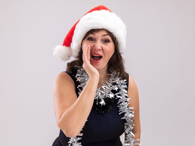 Impressionné femme d'âge moyen portant bonnet de noel et guirlande de guirlandes autour du cou regardant la caméra chuchoter isolé sur fond blanc avec espace de copie