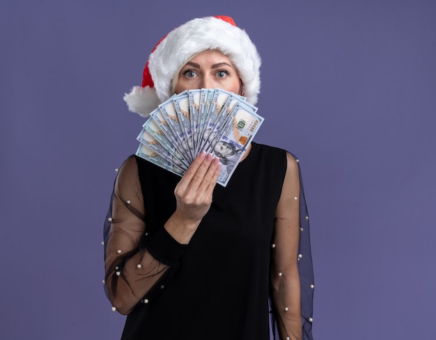 Impressionné femme blonde d'âge moyen portant un chapeau de Noël tenant de l'argent par derrière isolé sur mur violet avec espace copie