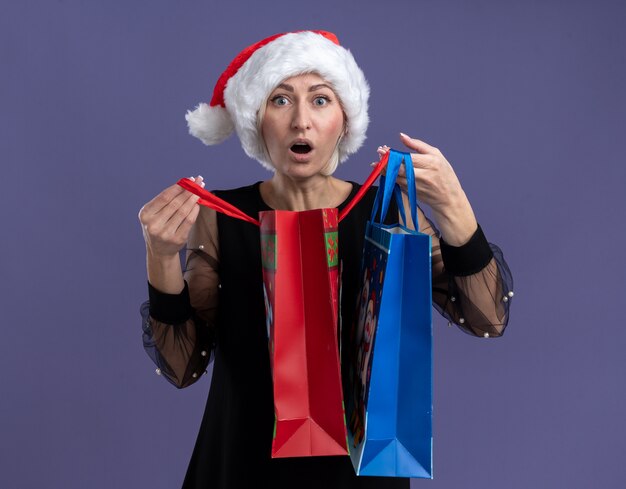 Impressionné femme blonde d'âge moyen portant un chapeau de Noël tenant des sacs-cadeaux de Noël ouverture d'un regardant la caméra isolée sur fond violet