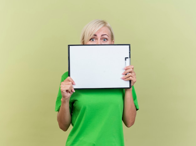 Impressionné femme blonde d'âge moyen tenant le presse-papiers en face de la bouche à l'avant par derrière il isolé sur mur vert olive