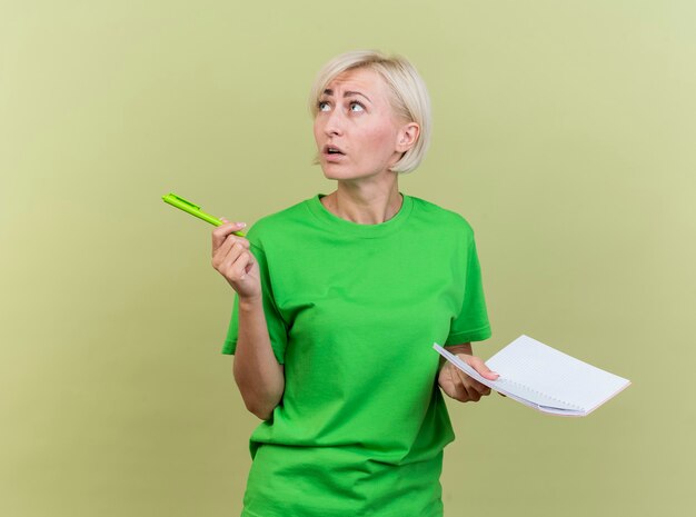 Impressionné femme blonde d'âge moyen tenant un stylo et un bloc-notes à côté isolé sur mur vert olive