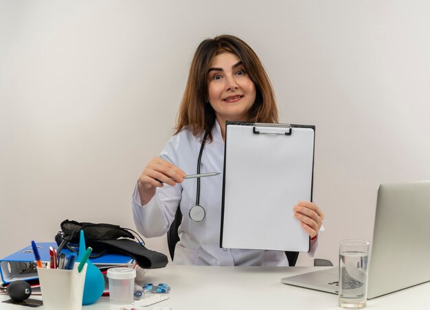 Impressionné femme médecin d'âge moyen portant une robe médicale et un stéthoscope assis au bureau avec des outils médicaux et un ordinateur portable tenant et pointant avec un stylo au presse-papiers isolé