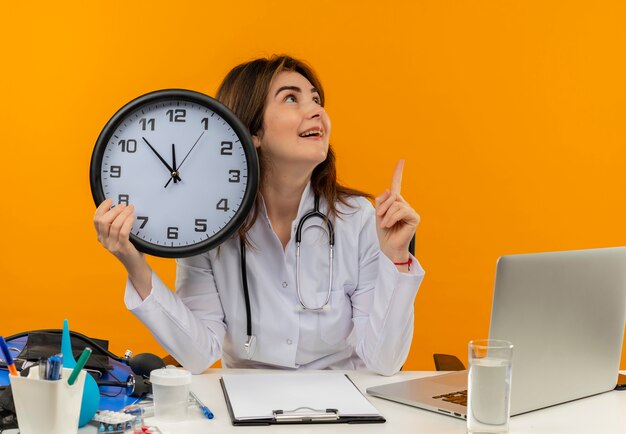 Impressionné femme médecin d'âge moyen portant une robe médicale et un stéthoscope assis au bureau avec presse-papiers d'outils médicaux et ordinateur portable tenant horloge regardant côté pointant vers le haut isolé