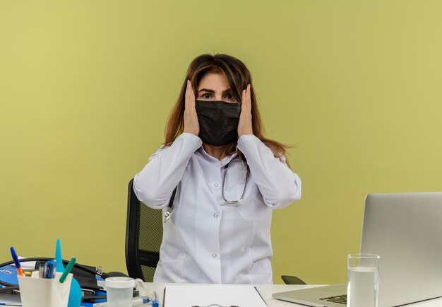 Impressionné femme médecin d'âge moyen portant une robe médicale et un stéthoscope et un masque assis au bureau avec des outils médicaux et un ordinateur portable mettant les mains sur la tête isolée