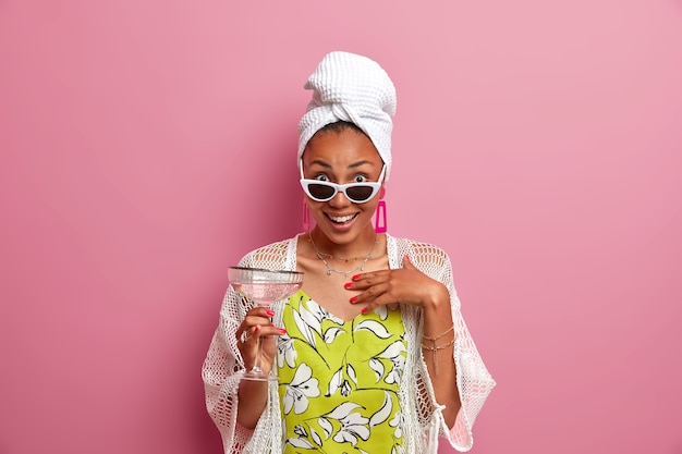 Impressionné heureux modèle féminin ethnique à la peau sombre tient un cocktail alcoolisé, porte des lunettes de soleil élégantes, une serviette de bain enveloppée sur la tête, passe du temps libre à une soirée pyjama, pose contre un mur rose